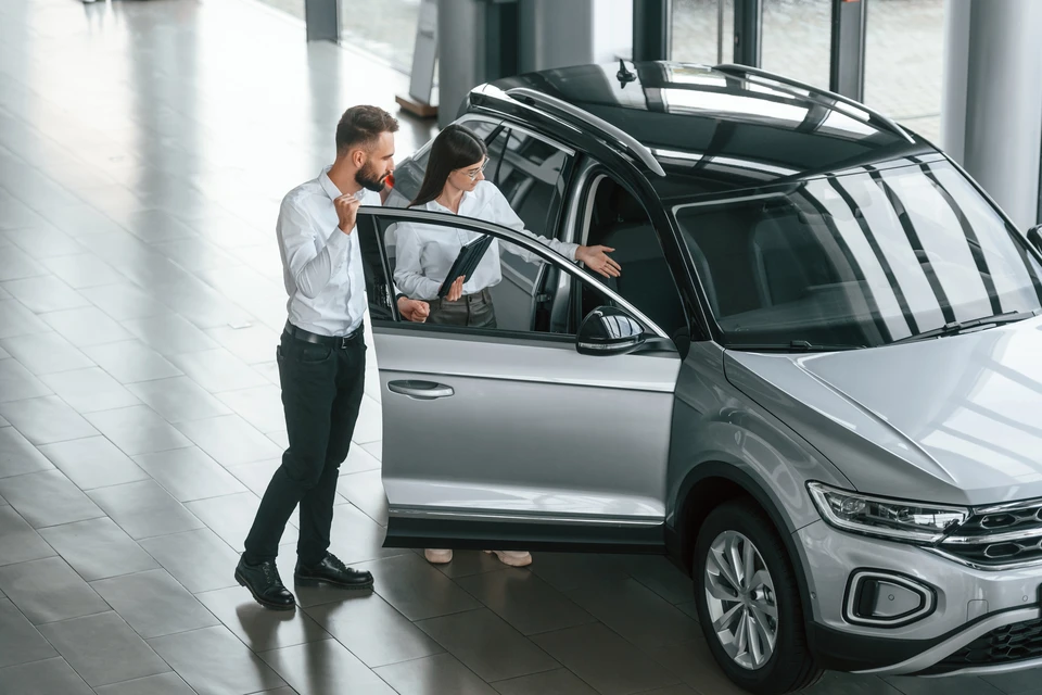 Concession automobile moderne avec vendeur en chemise blanche présentant SUV gris métallisé à cliente potentielle