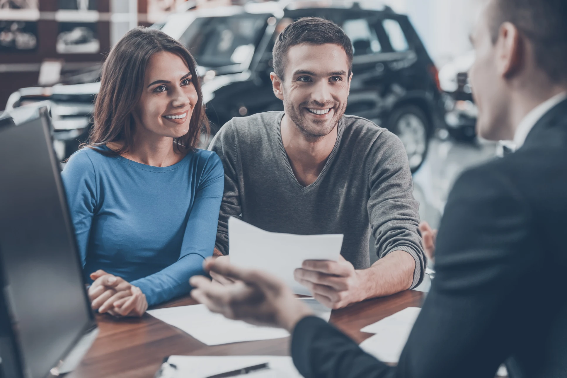 Couple souriant assis face à un conseiller dans un showroom automobile, examinant des documents contractuels sur une table en bois
