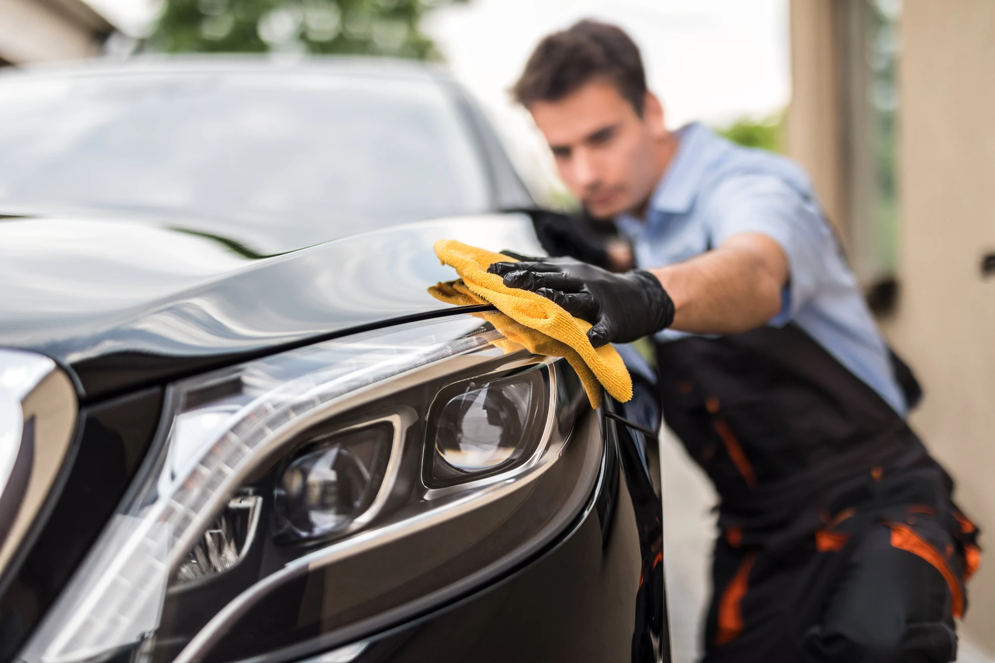 Un homme en bleu nettoie méticuleusement une voiture noire avec un chiffon jaune, portant des gants de protection